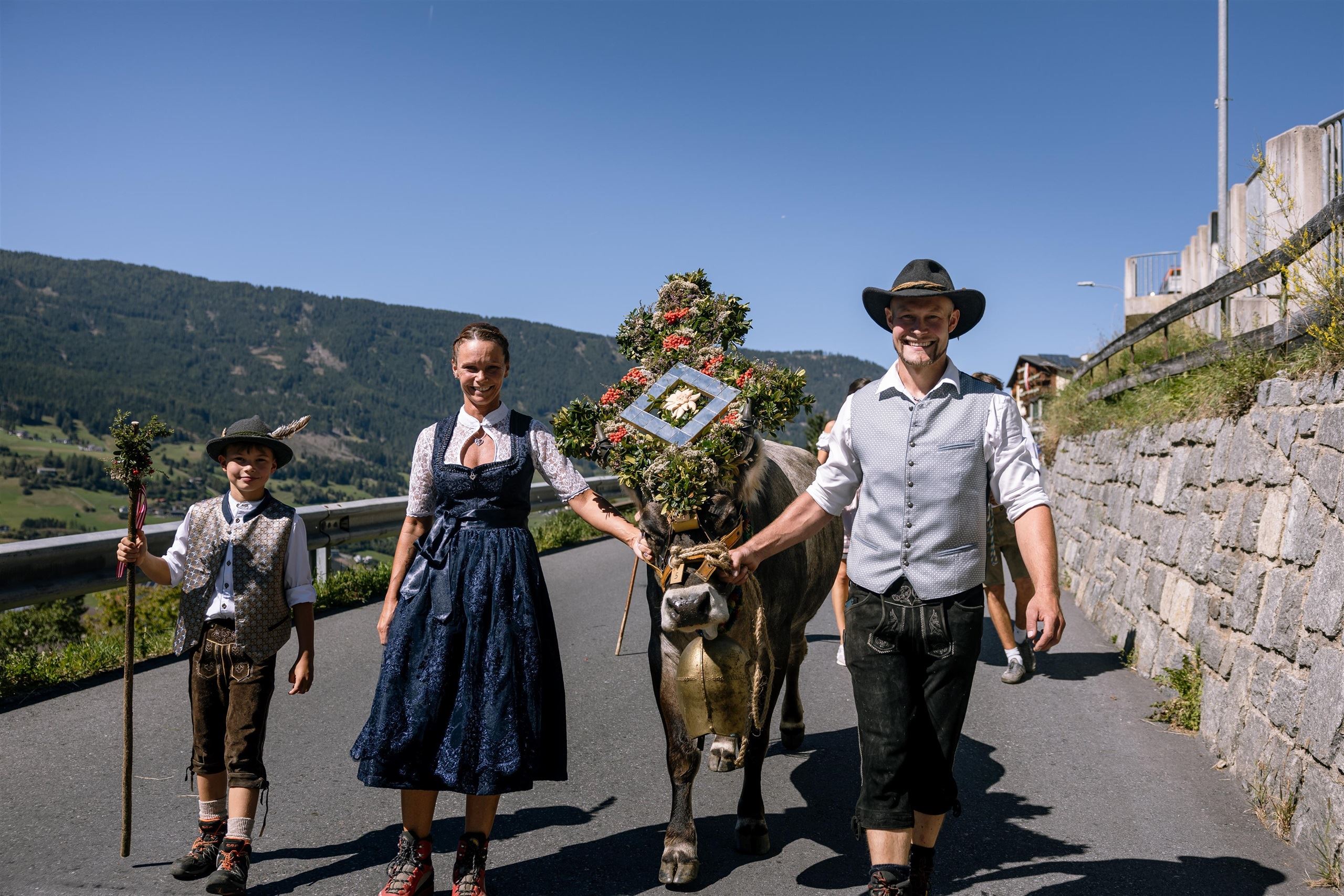 Eine Familie in traditioneller Tracht führt eine dekorierte Kuh auf einem landwirtschaftlichen Weg. Im Hintergrund sind Berge und ein blauer Himmel zu sehen.