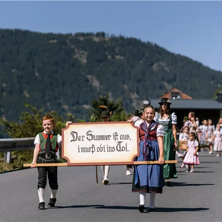 Eine Gruppe von Menschen in traditioneller Kleidung marschiert eine Straße entlang. Sie tragen ein Banner mit der Aufschrift: "Der Sommer ist aus, im nehm ich wohl ins Tal."