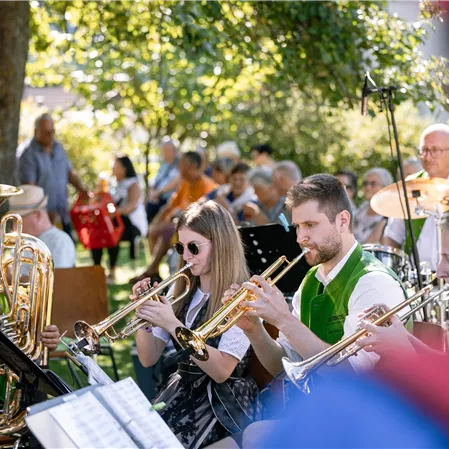 Eine lebhafte Musikgruppe spielt im Freien unter Bäumen. Im Hintergrund sind zahlreiche Zuhörer zu sehen, die die Darbietung genießen.