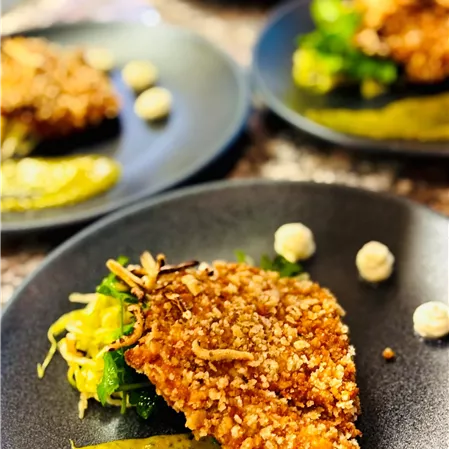 A deliciously arranged dish on a black plate. It features breaded fish with side dishes of vegetables and a sauce.
