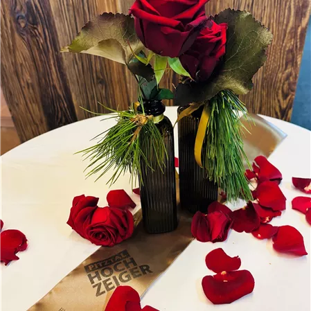 An elegant table with two roses in a decorative vase and scattered red rose petals. In the background lies a golden fabric with an inscription.