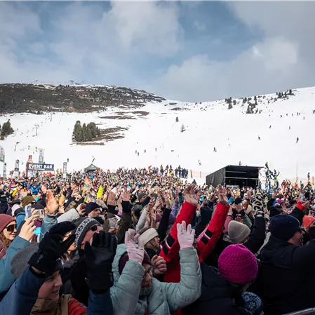 Eine große Menschenmenge feiert in einem verschneiten Skigebiet. Im Hintergrund sind schneebedeckte Berge und Skifahrer zu sehen.