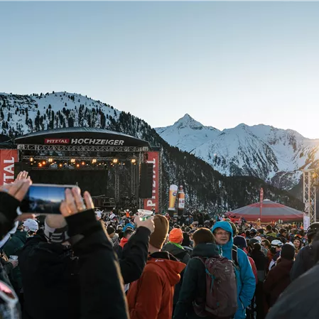 Ein Konzert vor schneebedeckten Bergen mit einer großen Menschenmenge. Die Sonne scheint im Hintergrund und sorgt für eine festliche Atmosphäre.