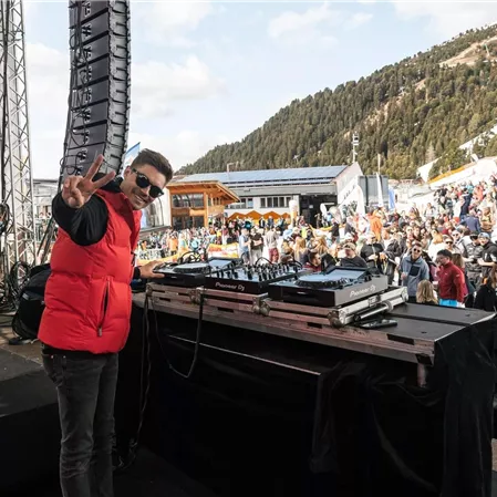 Ein DJ steht an einem Mischpult und gibt ein Zeichen der Freude. Im Hintergrund ist eine große Menschenmenge auf einer Veranstaltung in den Bergen zu sehen.