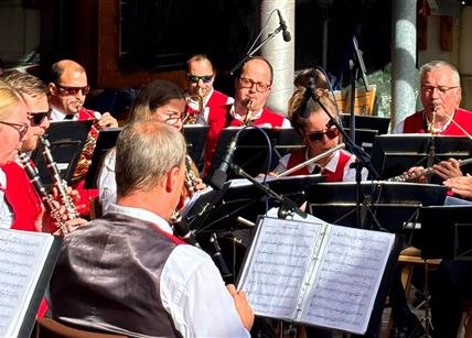 Eine Blaskapelle spielt in einem Freiluftkonzert. Die Musiker tragen rote Westen und konzentrieren sich auf die Aufführung.