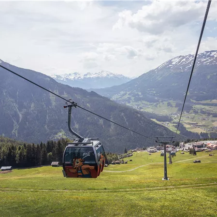Eine Seilbahn fährt über grüne Wiesen in den Bergen. Im Hintergrund sind schneebedeckte Gipfel und ein idyllisches Tal zu sehen.