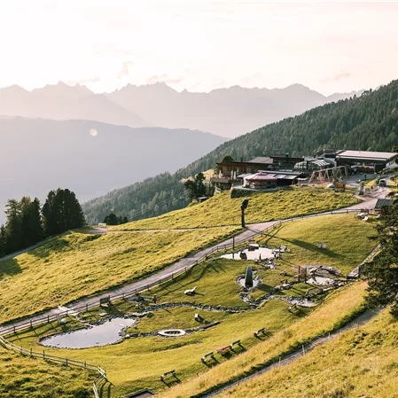 Eine malerische Landschaft mit sanften Hügeln und grünen Wiesen. Im Hintergrund sind Berge und ein Gebäude sichtbar, das in die Natur integriert ist.