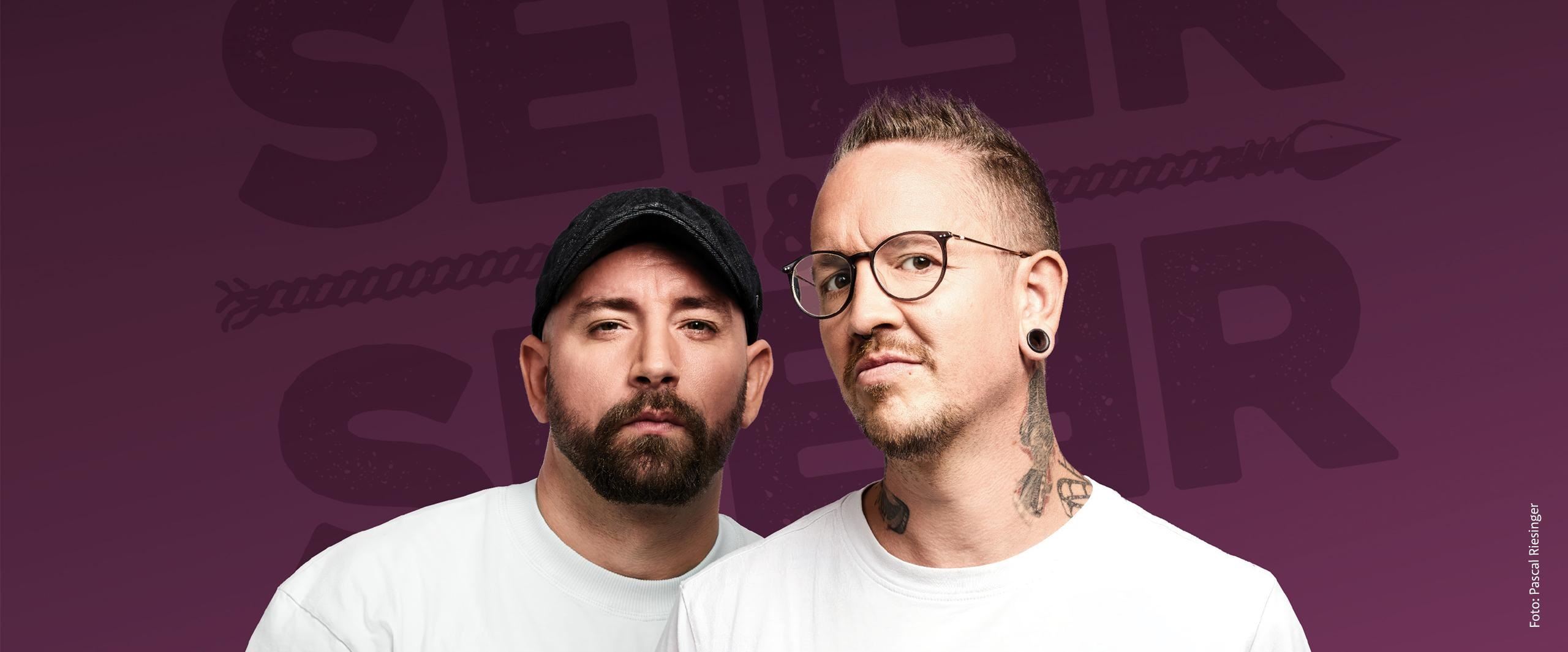 Zwei Männer stehen vor einem lila Hintergrund und schauen in die Kamera. Sie tragen weiße T-Shirts und haben jeweils verschiedene Stile, darunter Tattoos und eine Mütze.