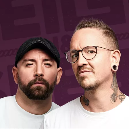 Zwei Männer stehen vor einem lila Hintergrund und schauen in die Kamera. Sie tragen weiße T-Shirts und haben jeweils verschiedene Stile, darunter Tattoos und eine Mütze.