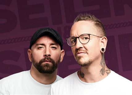 Zwei Männer stehen vor einem lila Hintergrund und schauen in die Kamera. Sie tragen weiße T-Shirts und haben jeweils verschiedene Stile, darunter Tattoos und eine Mütze.