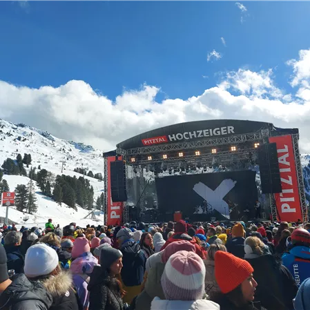 Eine große Menschenmenge steht vor einer Bühne in den Bergen, umgeben von Schnee. Der Himmel ist blau mit einigen Wolken.