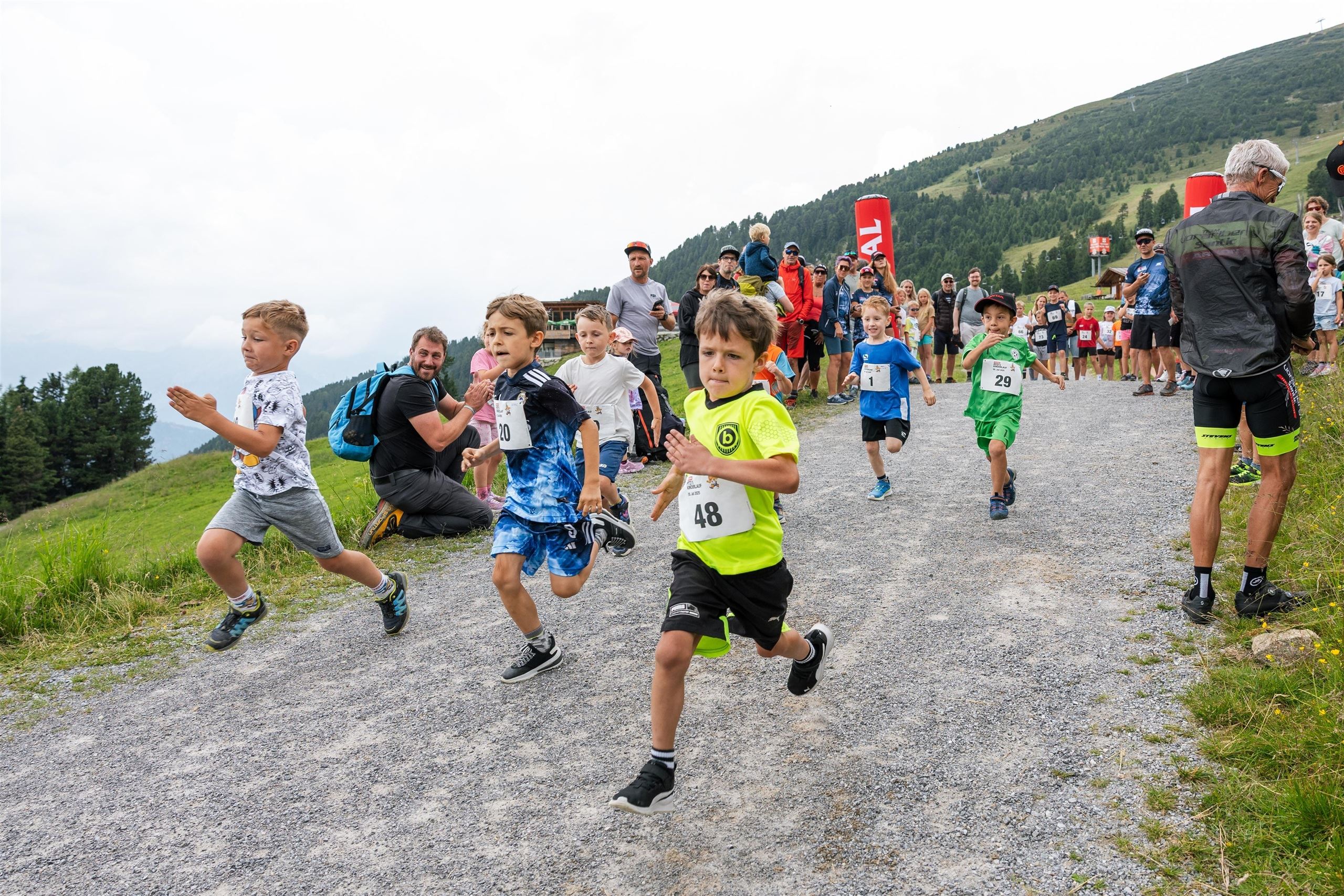 Eine Gruppe von Kindern läuft enthusiastisch einen Feldweg entlang. Im Hintergrund sind Zuschauer und eine hügelige Landschaft zu sehen.