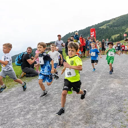 Eine Gruppe von Kindern läuft enthusiastisch einen Feldweg entlang. Im Hintergrund sind Zuschauer und eine hügelige Landschaft zu sehen.