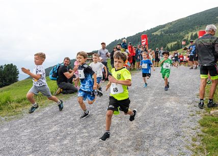 Eine Gruppe von Kindern läuft enthusiastisch einen Feldweg entlang. Im Hintergrund sind Zuschauer und eine hügelige Landschaft zu sehen.