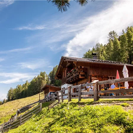 Ein traditionelles Chalet in den Bergen, umgeben von grünen Wiesen und Bäumen. Der Himmel ist blau mit weißen Wolken, und es sind einige Menschen in der Nähe.