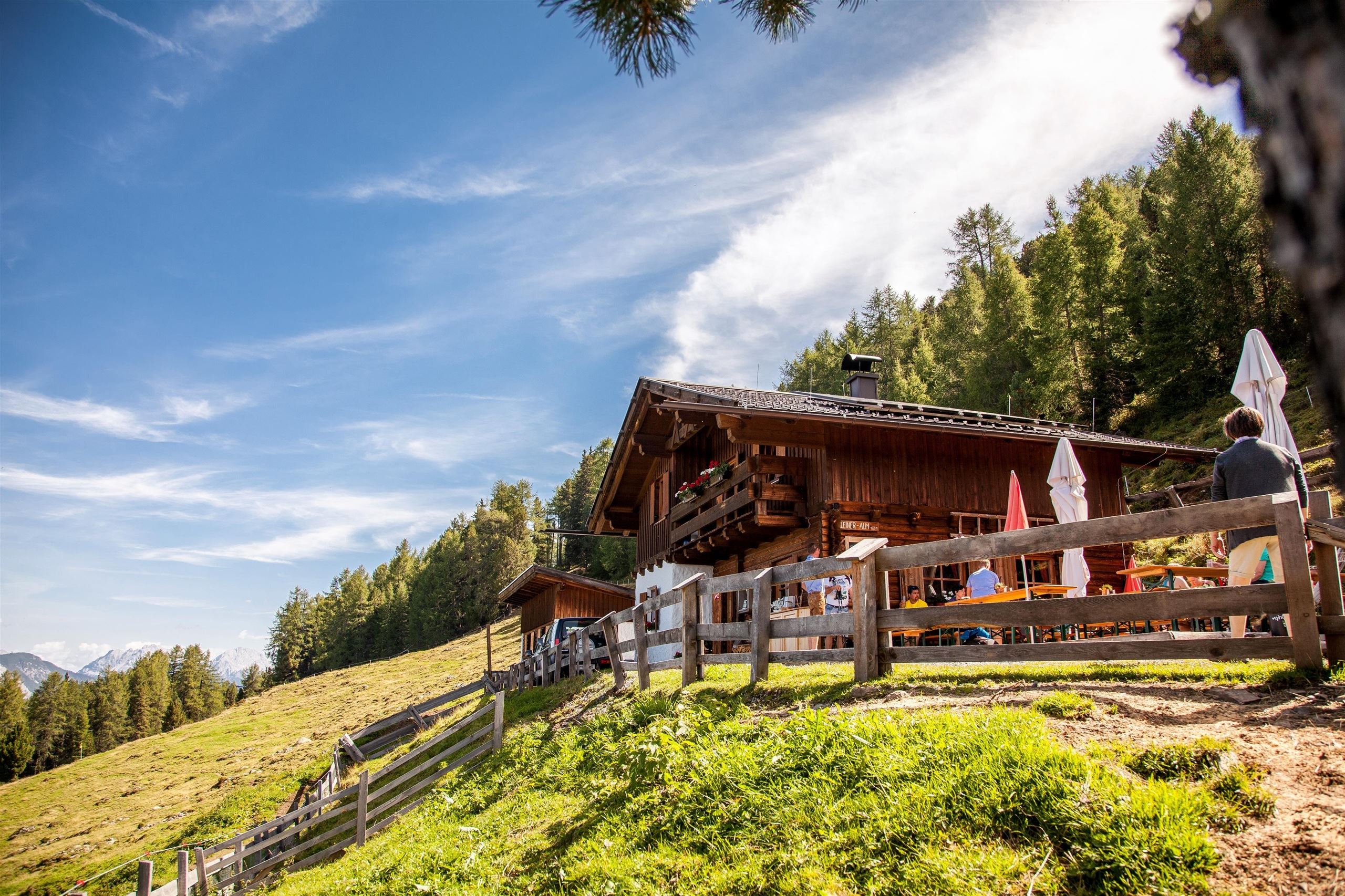 Ein traditionelles Chalet in den Bergen, umgeben von grünen Wiesen und Bäumen. Der Himmel ist blau mit weißen Wolken, und es sind einige Menschen in der Nähe.