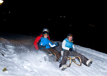 Zwei Personen fahren mit dem Schlitten die verschneite Piste hinunter. Es ist Nacht, und etwas Licht erhellt die Szene.