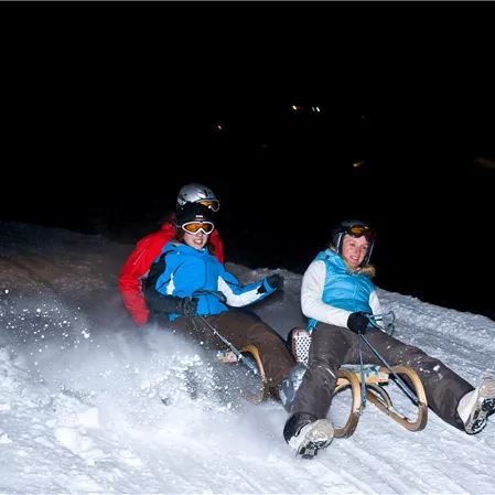 Zwei Personen fahren mit dem Schlitten die verschneite Piste hinunter. Es ist Nacht, und etwas Licht erhellt die Szene.