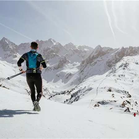 Ein Mann läuft auf einer schneebedeckten Berglandschaft. Im Hintergrund sind majestätische Berge und ein klarer blauer Himmel zu sehen.