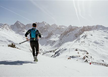 Ein Mann läuft auf einer schneebedeckten Berglandschaft. Im Hintergrund sind majestätische Berge und ein klarer blauer Himmel zu sehen.
