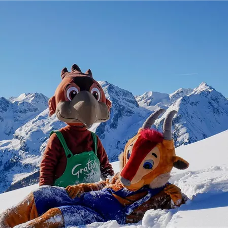 Zwei Maskottchen stehen im Schnee vor einer beeindruckenden Berglandschaft. Der Himmel ist klar und blau, während die schneebedeckten Berge im Hintergrund sichtbar sind.