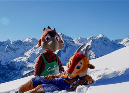 Zwei Maskottchen stehen im Schnee vor einer beeindruckenden Berglandschaft. Der Himmel ist klar und blau, während die schneebedeckten Berge im Hintergrund sichtbar sind.