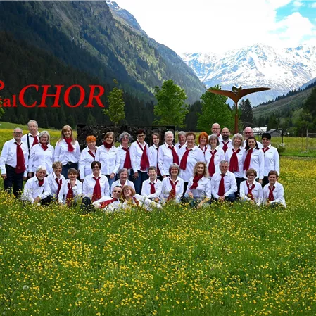 Eine Gruppe von Sängern steht in einem blühenden Feld vor einer beeindruckenden Berglandschaft. Alle tragen elegante Kleidung in Weiß und Rot.