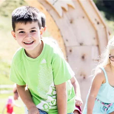Zwei Kinder spielen draußen auf einem Spielplatz. Im Hintergrund sind grüne Wiesen und Bäume sichtbar.