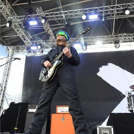 Ein Musiker spielt Gitarre auf einer Bühne. Er trägt eine schwarze Kleidung und Sonnenbrille.
