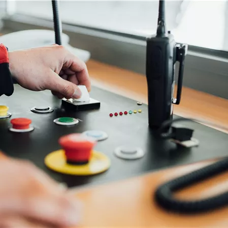 A hand operates a control panel with various buttons in different colors. In the background, a radio is visible.