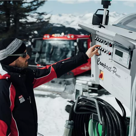 A man operates a snow machine in a snowy landscape. In the background, there are mountains and a groomer.