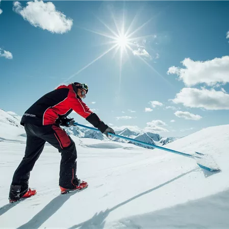 A person is shoveling snow in a sunny mountain landscape. The sky is blue and covered with some clouds.