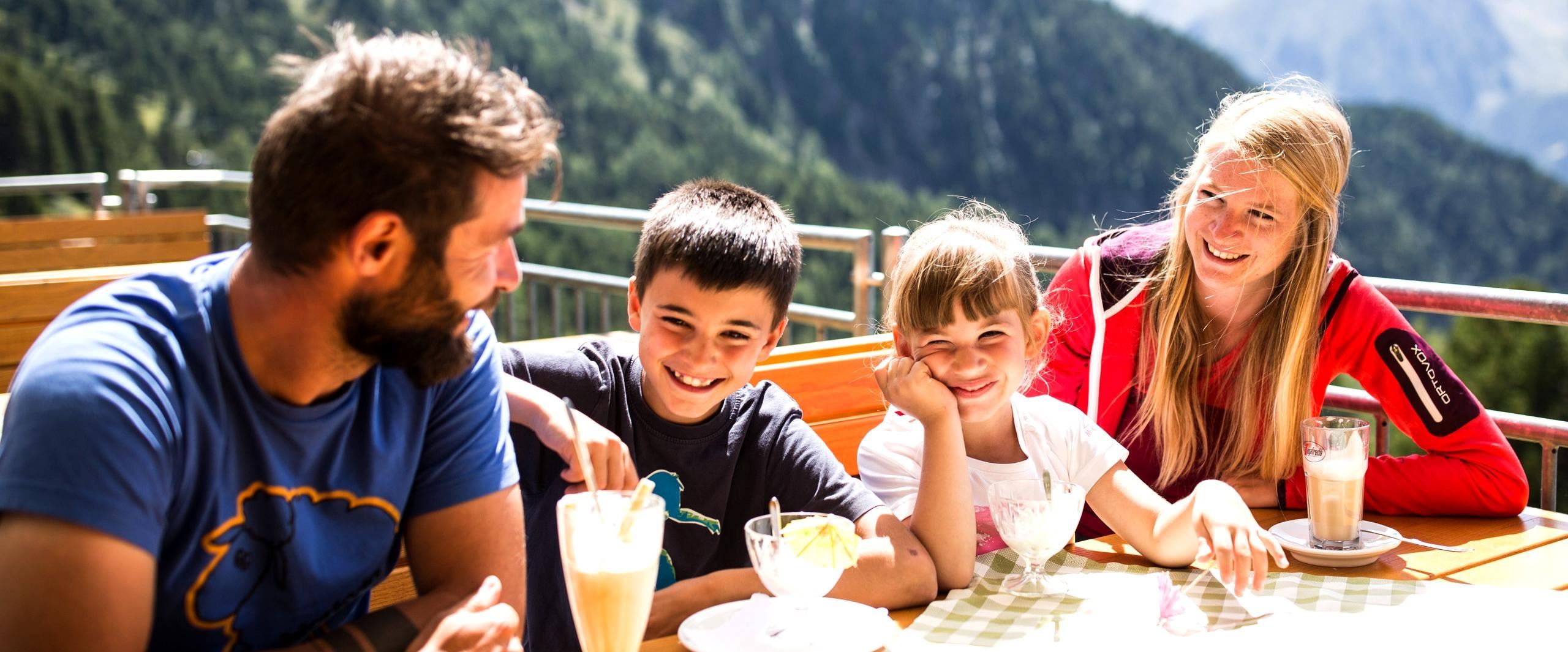 Eine fröhliche Familie sitzt an einem Tisch im Freien und genießt Eis und Getränke. Im Hintergrund sind Berge und eine grüne Landschaft zu sehen.