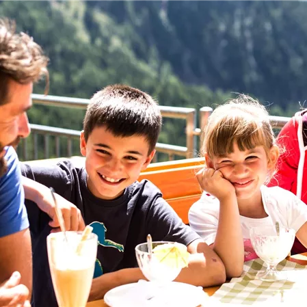 Eine fröhliche Familie sitzt an einem Tisch im Freien und genießt Eis und Getränke. Im Hintergrund sind Berge und eine grüne Landschaft zu sehen.