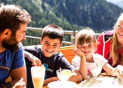 Eine fröhliche Familie sitzt an einem Tisch im Freien und genießt Eis und Getränke. Im Hintergrund sind Berge und eine grüne Landschaft zu sehen.