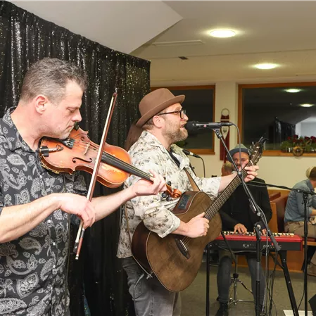 A live band is playing in a cozy room. Two musicians are in the foreground, one with a violin and the other with a guitar.