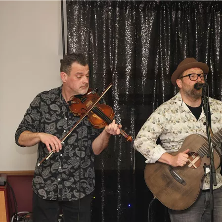 Two musicians are performing on a stage. The man on the left plays the violin, while the man on the right plays the guitar and sings.