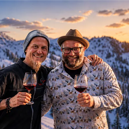 Two men are standing in the snow holding a glass of red wine in their hands. In the background, the sun is shining over snow-covered mountains.
