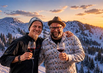 Two men are standing in the snow holding a glass of red wine in their hands. In the background, the sun is shining over snow-covered mountains.
